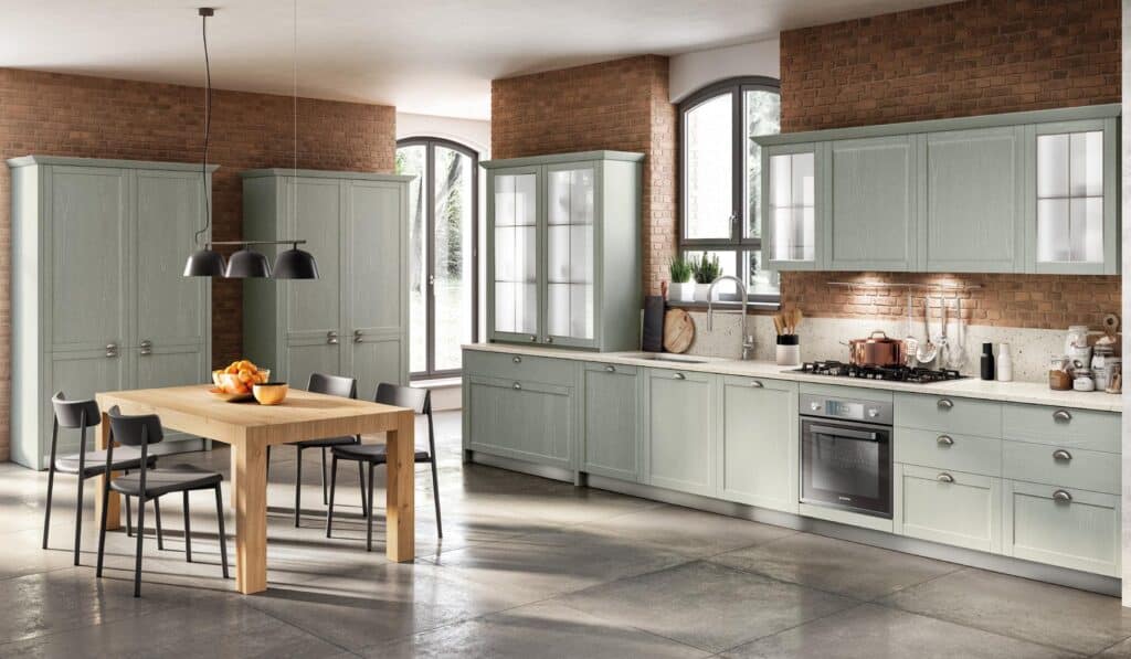 Modern kitchen with light green cabinets, a wooden dining table with black chairs, brick walls, large windows, and stainless steel appliances.