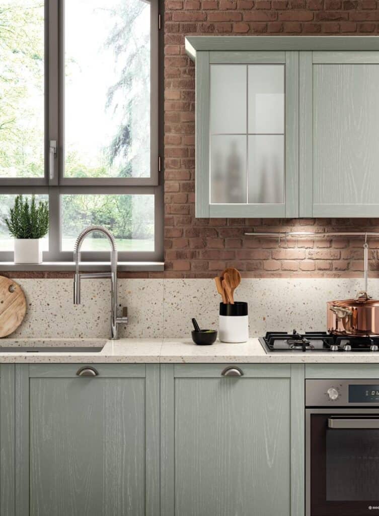 Modern kitchen with light green cabinets, a terrazzo countertop and backsplash, and a brick wall. Features include a metal faucet, a gas stove, and a window with outdoor greenery visible.