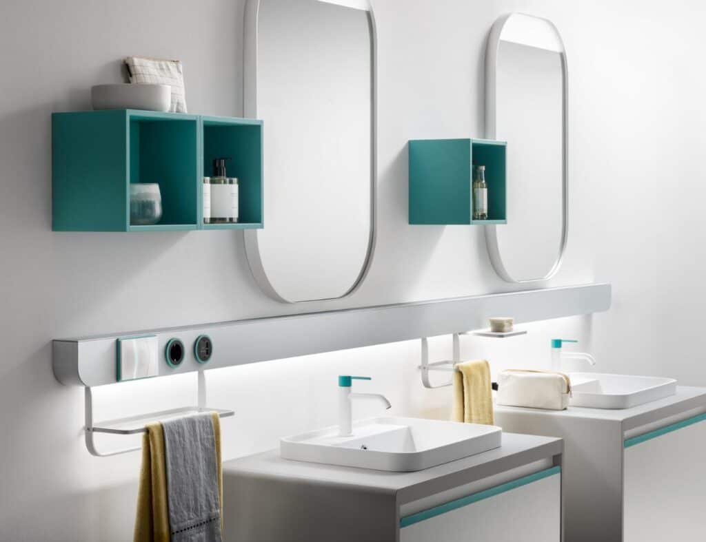 Modern bathroom with teal wall shelves, oval mirrors, and two sinks with white countertops and yellow towels.
