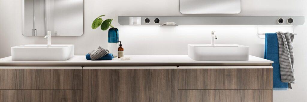 Modern bathroom vanity with two sinks, a wooden cabinet, wall mirrors, and minimalist decor including towels, a plant, and toiletries.