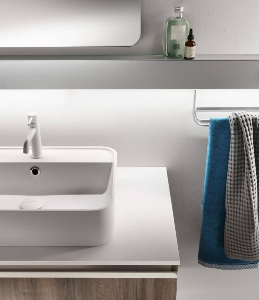 Modern bathroom sink with a white rectangular basin, a white faucet, a glass shelf above holding bottles, and a towel rail with blue and gray towels.