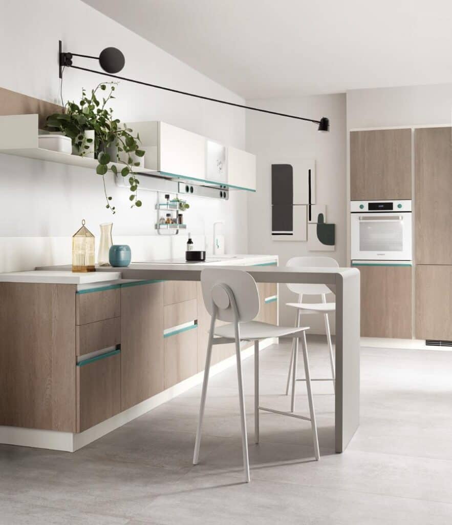 Modern kitchen with light wood cabinets, a white island, minimalist bar stools, and a wall-mounted lamp. A potted plant and decorative items adorn the space.