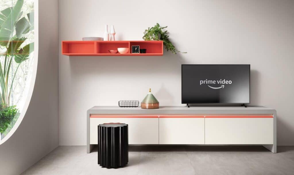 Modern living room with a wall-mounted TV displaying &quot;prime video,&quot; a coral shelf with decor items, a white and coral cabinet, a black stool, and a large window with plants.