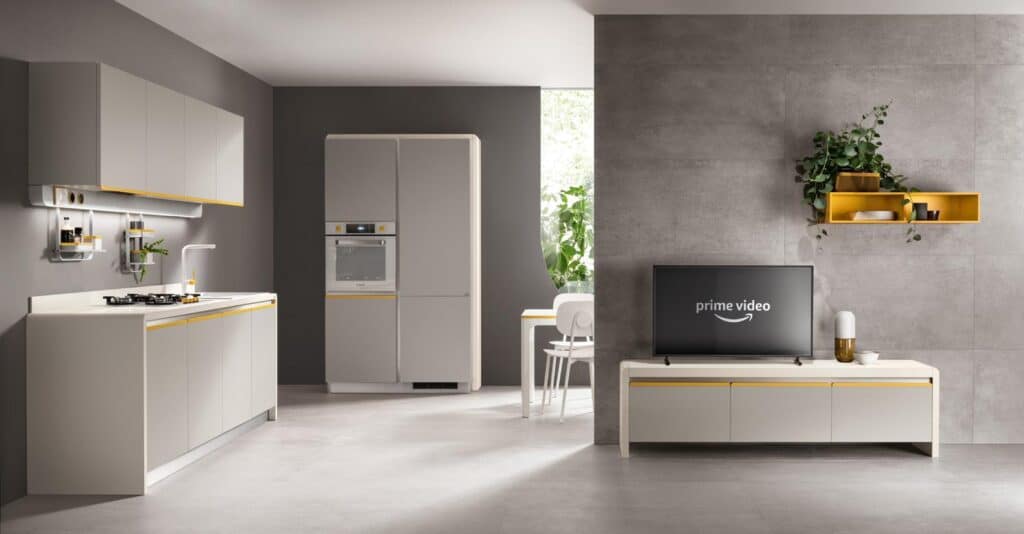 Modern kitchen and living area with minimalist design, featuring gray cabinets, a wall-mounted TV, and open shelving with plants and decor against a concrete backdrop.
