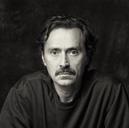 Black and white portrait of a man with a mustache wearing a dark sweater, looking directly at the camera.