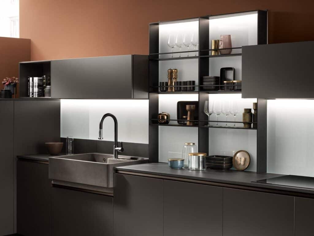 Modern kitchen interior with sleek dark cabinetry, open shelving displaying glassware and decor, and a large metal sink under a tall faucet.