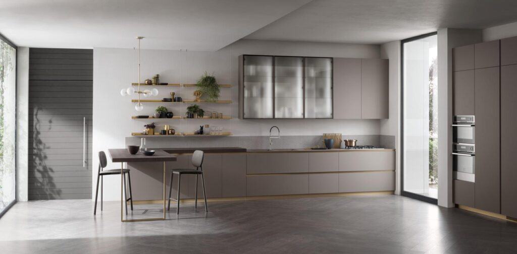 Modern kitchen with sleek gray cabinetry, open shelves with plants and decor, a large countertop, built-in appliances, pendant lighting, and a chaise lounge by the window.