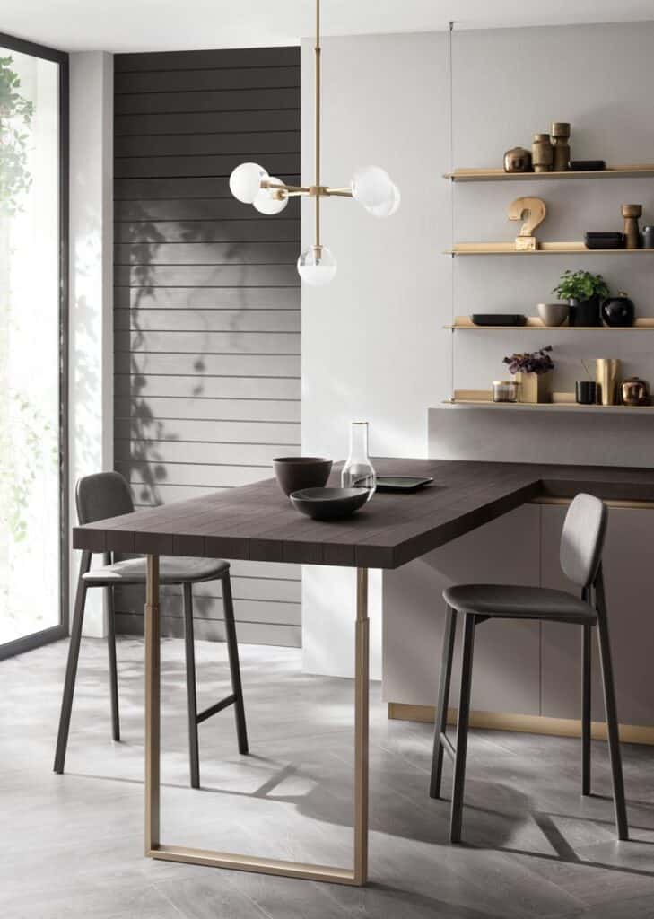 Modern kitchen with a dark wood table, grey bar stools, and minimalist shelves with decor against light grey walls; a geometric light fixture hangs above.