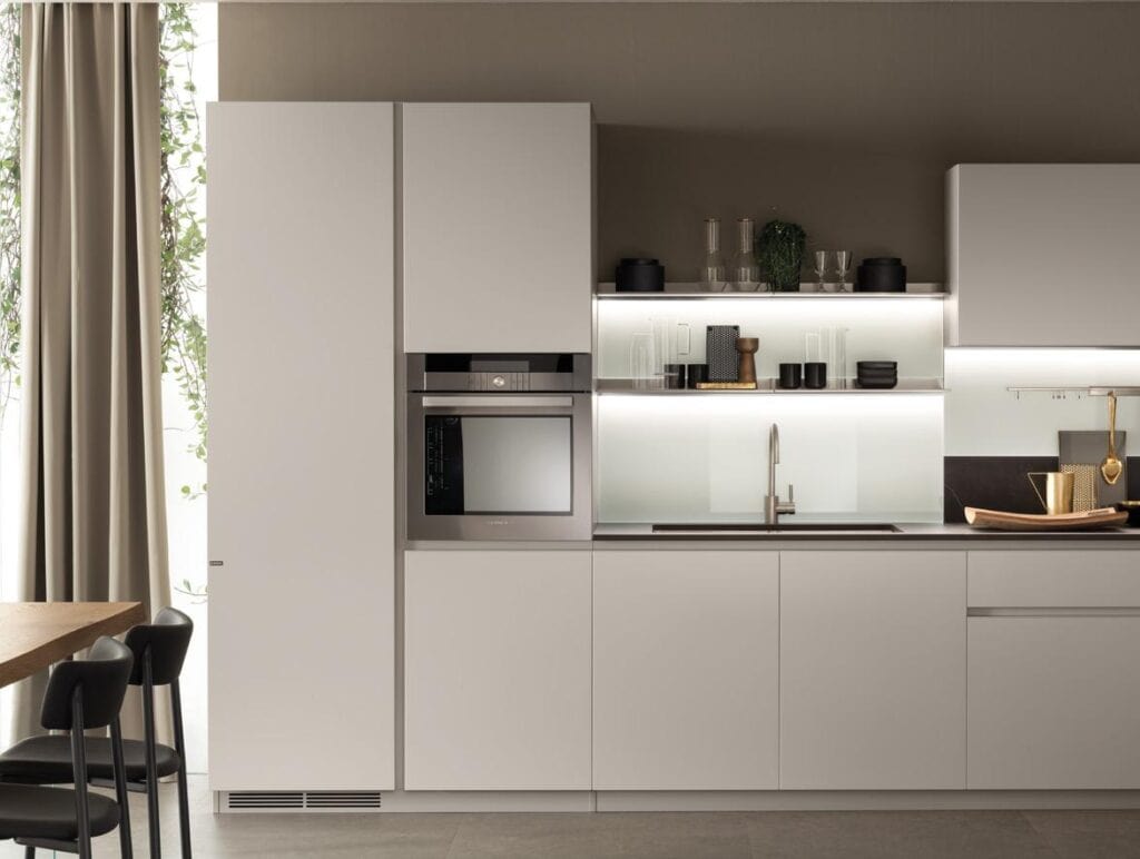 Modern kitchen with white cabinetry, built-in oven, open shelving with glassware and decor, and a sink under a lit backsplash.