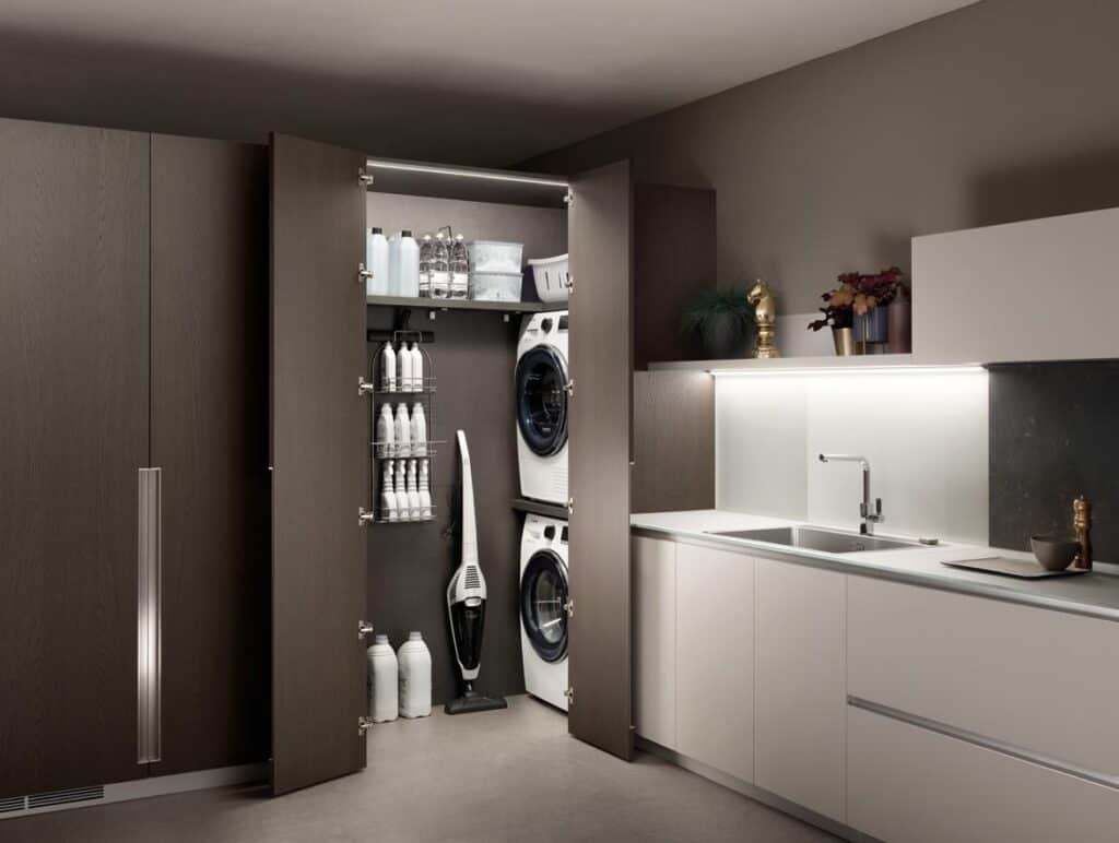 Modern kitchen with open cupboard revealing a stacked washer and dryer, cleaning supplies, and a vacuum cleaner.