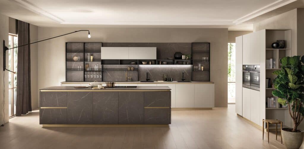 Modern kitchen with gray and beige cabinetry, a large island, open shelving, built-in ovens, and a potted plant.