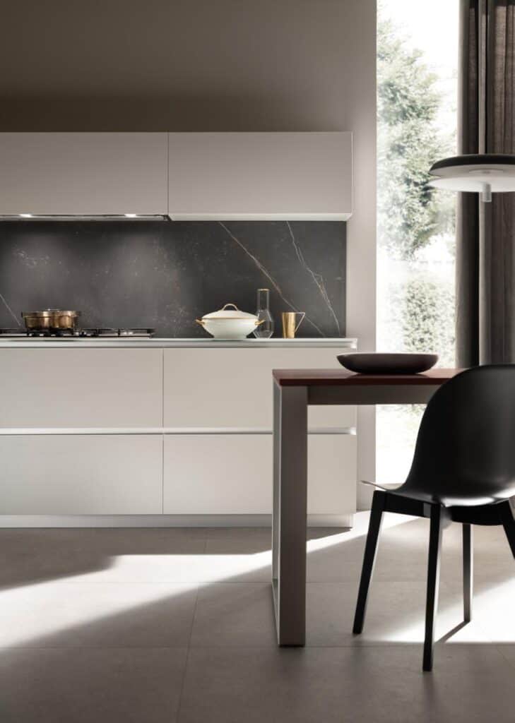 Modern kitchen interior with minimalist white cabinetry, dark backsplash, black chair, and a tabletop with a bowl, under natural light.