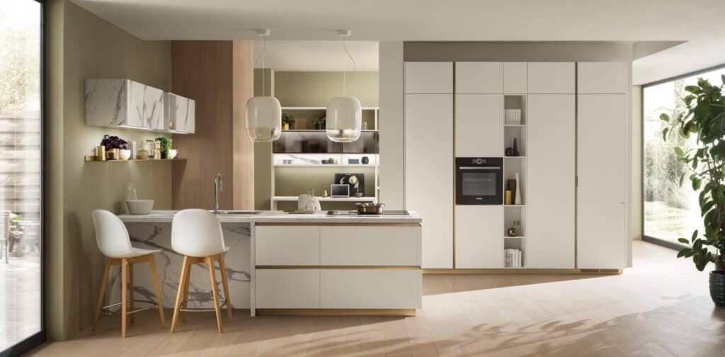 Modern kitchen with marble countertops, white cabinetry, and built-in appliances, featuring a central island with barstools and pendant lighting, set in a room with large windows and wooden flooring.