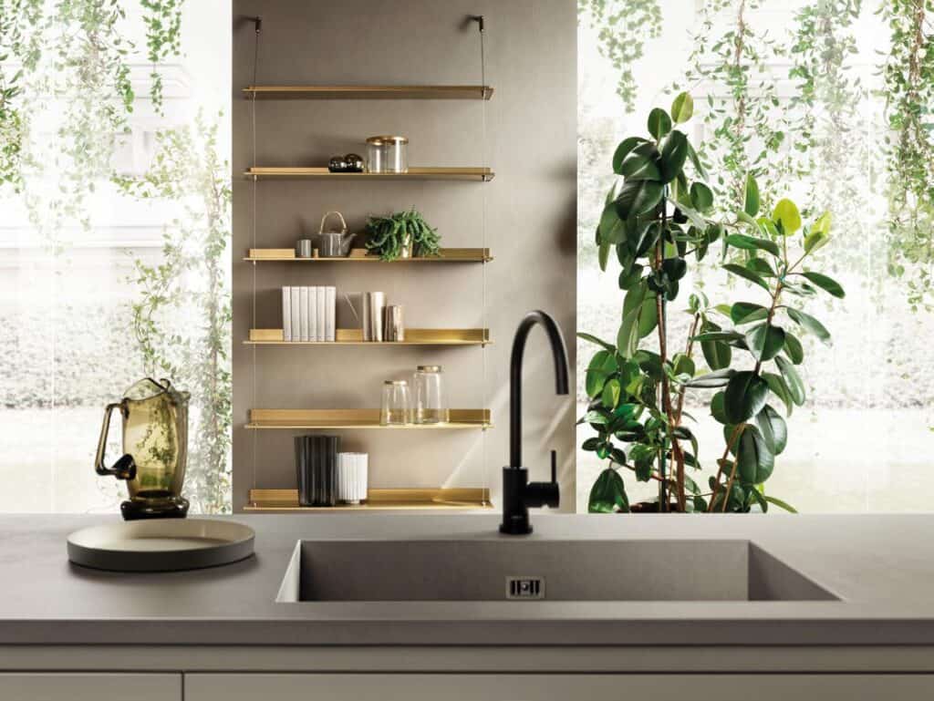Modern kitchen interior with a black faucet, sleek countertop, golden shelves holding books and glassware, and lush green plants.