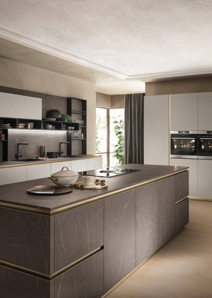 Modern kitchen with a large island featuring dark marble-like surfaces, a cooktop, and various kitchen items. Light cabinets and an oven are visible in the background, with open shelving and a window with curtains.