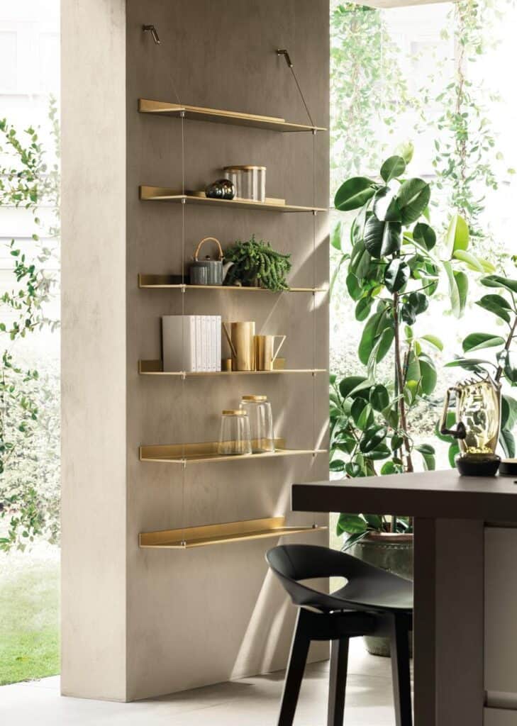 Delinea-3 - Inspo Living Spaces Modern wall-mounted shelves with golden accents holding decorative items in a bright room with large plants.
