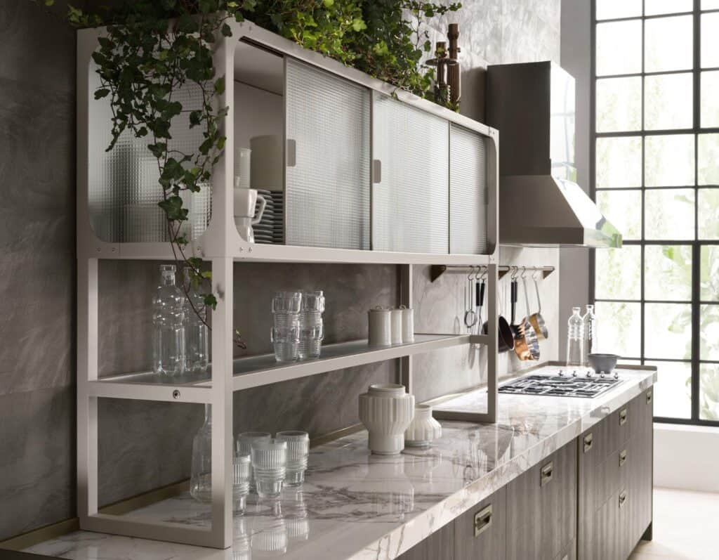 Modern kitchen interior with marble countertops, open shelving with dishes, glassware, and green plants, and a stove with hanging utensils.