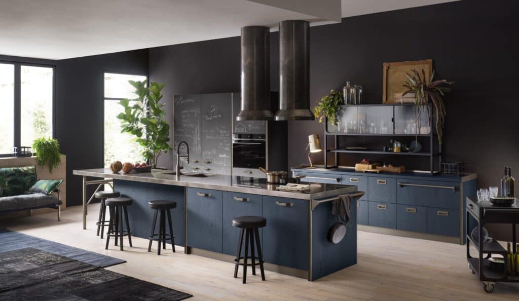 Modern kitchen with dark cabinetry, large island with stools, overhead metal range hood, chalkboard wall, and plants for decoration.