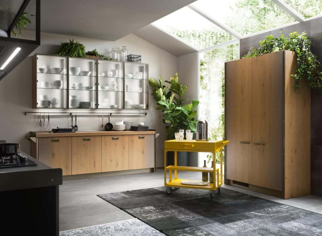 Modern kitchen with wooden cabinets, glass-front cupboards, yellow cart, black and white rug, and indoor plants under a skylight.