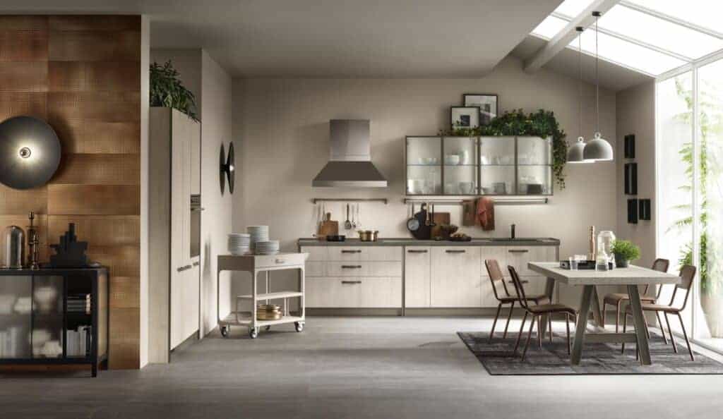 Modern kitchen with light wooden cabinets, a dining table with chairs, a serving cart, and pendant lights, featuring greenery and large windows.