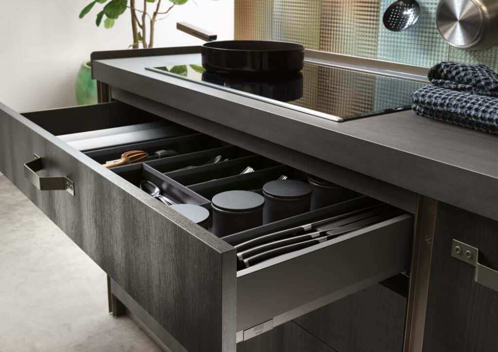 Open kitchen drawer with organized utensils, cutlery, and jars next to a stovetop.