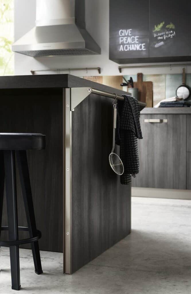 Modern kitchen interior with dark wood island, stainless steel extractor, black stool, and hanging kitchen towel and whisk; chalkboard with &quot;Give Peace a Chance&quot; written on it.