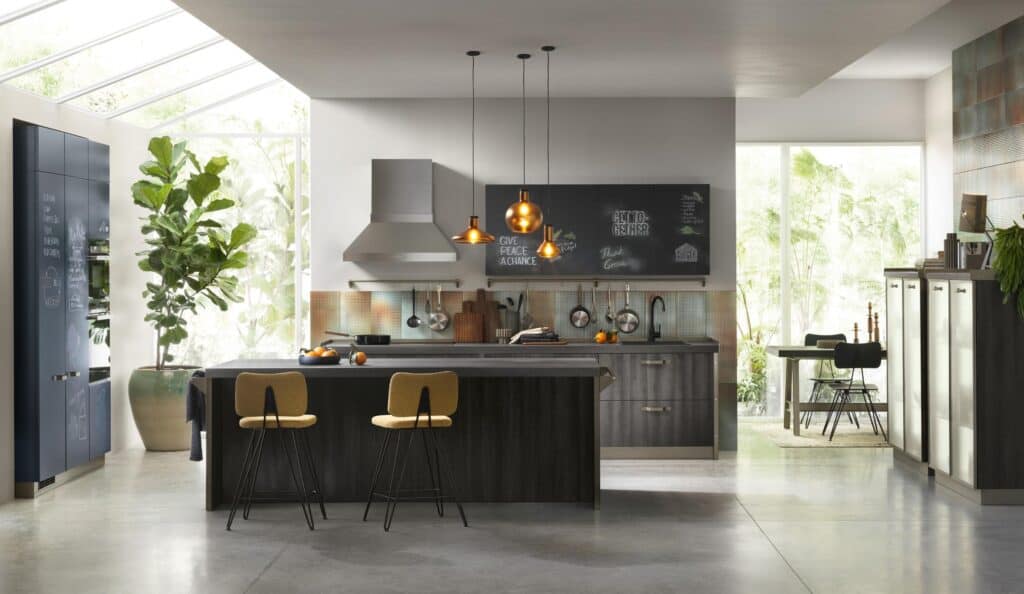 Modern kitchen with dark cabinetry, a large central island, pendant lights, and stools. A large potted plant is on the left, light streaming through skylights and large windows reveals a green garden outside.