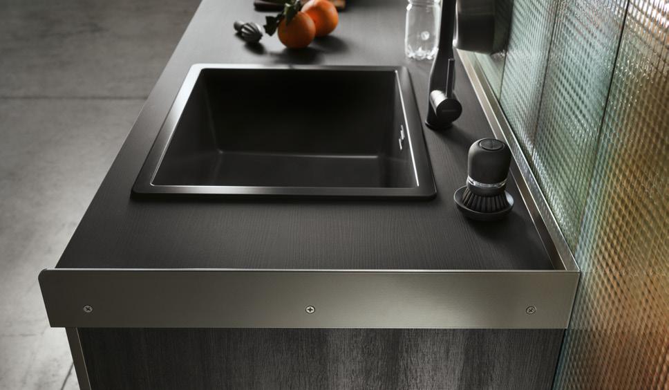 Modern kitchen countertop with a sleek black sink, faucet, and countertop brush, with oranges and a jar in the background.