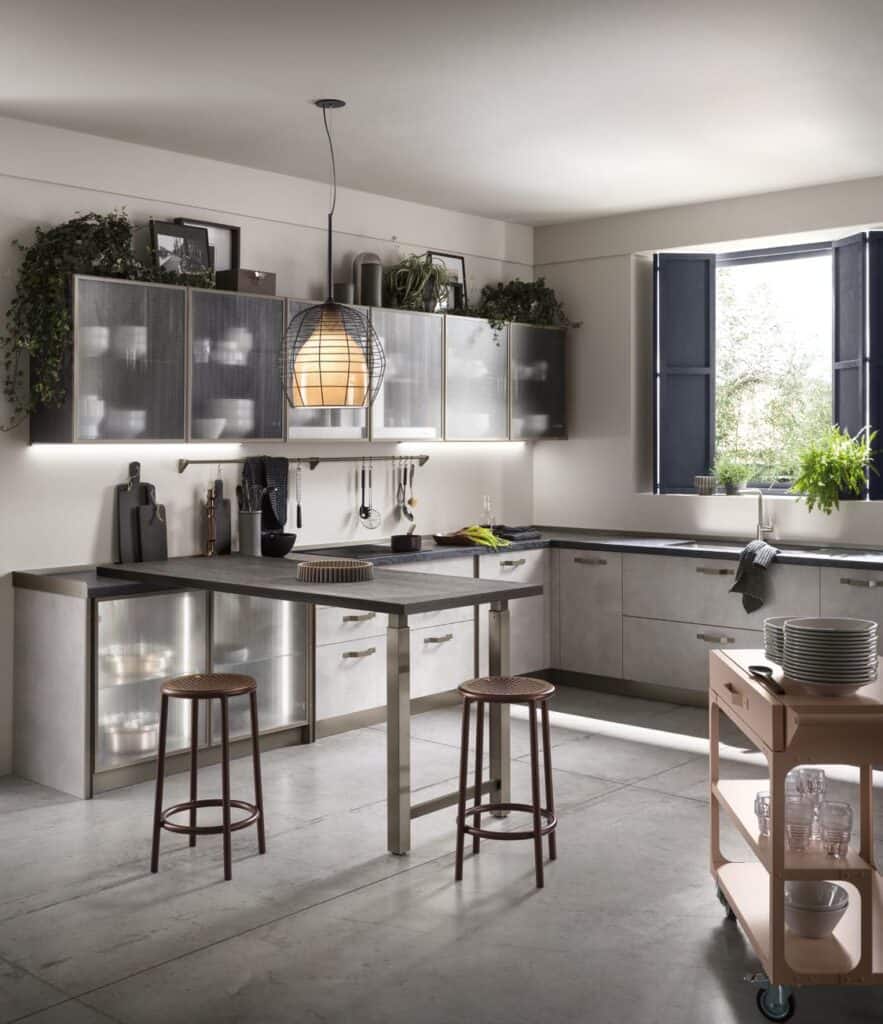 Modern kitchen with sleek gray cabinets, a central island with stools, a hanging pendant light, and a window with shutters. A rolling cart with dishes is in the foreground.