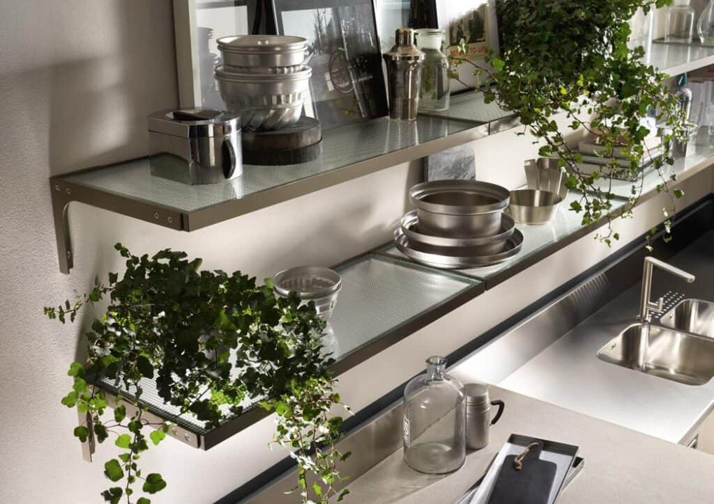 Kitchen shelves with metal cookware and glass decor, alongside leafy green plants, above a stainless steel sink.