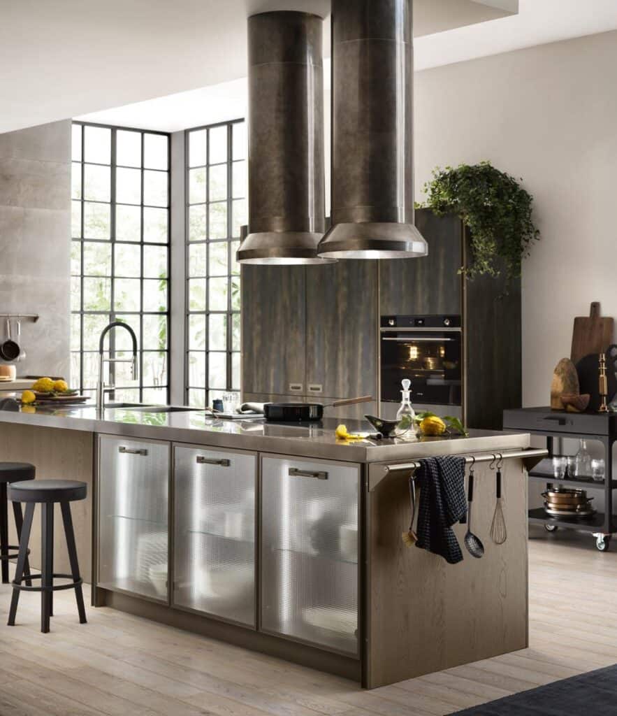 Modern kitchen with a large island, two metallic cylindrical range hoods, glass-front cabinets, and a black oven.