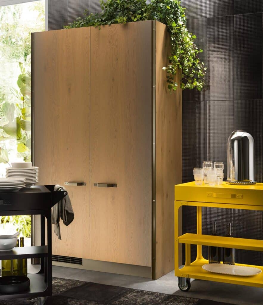 Wooden cabinet with greenery on top, accompanied by a yellow cart with glasses and a bottle, set against a dark textured wall.