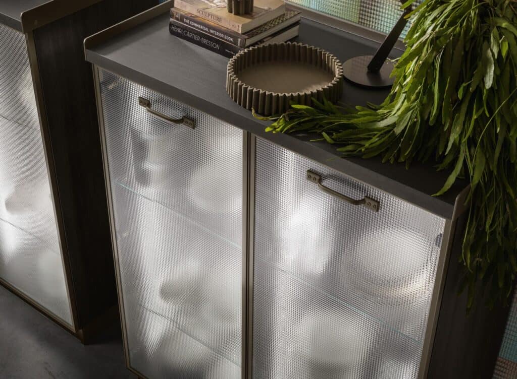 A dark wood cabinet with frosted glass doors, metal handles, and a gray top surface featuring books, a decorative bowl, and green foliage.