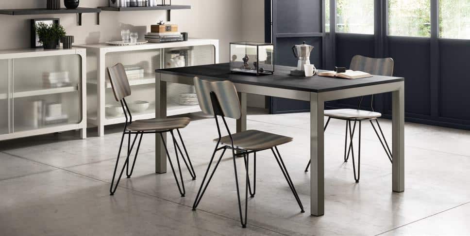 A modern dining area with a sleek table, four chairs, a coffee pot, and a book on the table. A cabinet is against the wall, and large windows provide natural light.