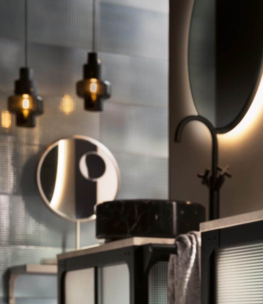 Blurry interior of a modern bathroom with hanging lights, a round mirror, a black faucet, and a textured countertop with a cloth.