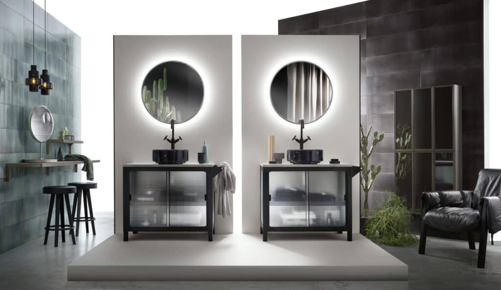 Modern bathroom interior with two illuminated circular mirrors, black sinks, and cabinets on a raised platform. A black leather chair and decorative cacti are visible in the background.