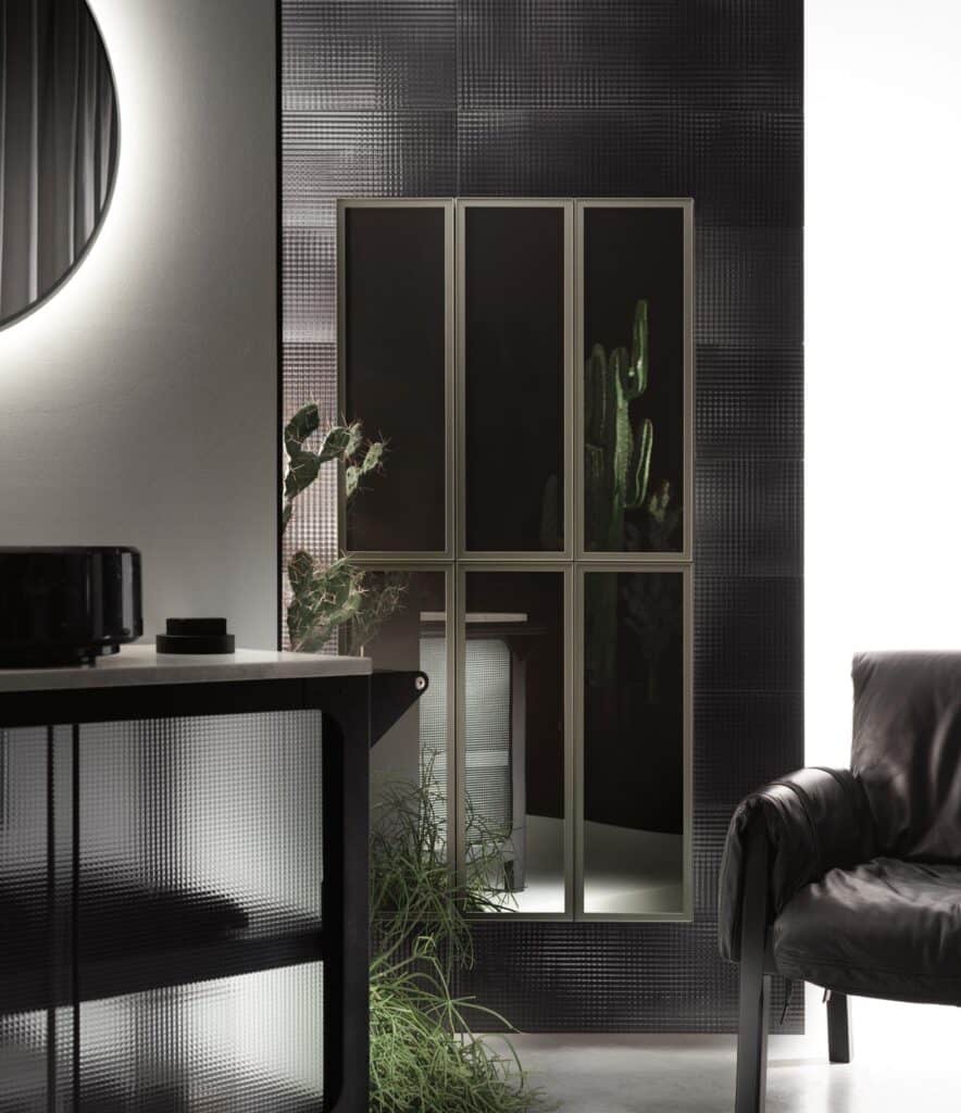 A modern interior featuring a textured black wall with a tall window, a circular mirror, a dark cabinet, and a black leather chair. There are green plants visible both inside and outside the window.