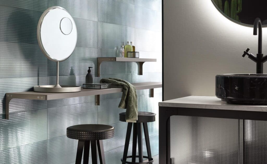 Modern bathroom with a large round mirror, glass panel wall, floating shelves with toiletries, and a black marble sink with a dark faucet.