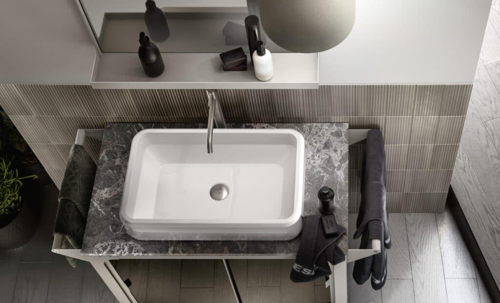 Modern bathroom with a rectangular white sink on a dark marble countertop, featuring a silver faucet, black and white toiletries, and towels.