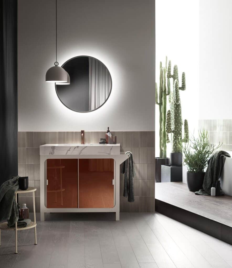 Modern bathroom interior with round mirror, hanging light fixture, white vanity with marble top, large potted cacti, and wood flooring.