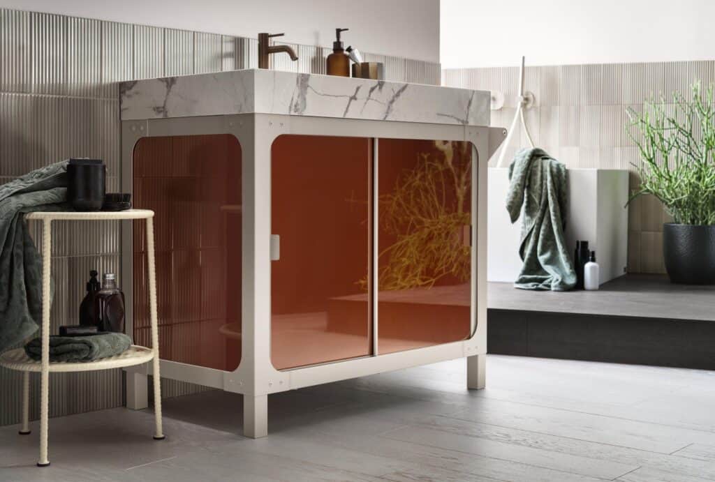 Modern bathroom vanity with a marble countertop and semi-transparent orange cabinet doors, accompanied by a small side table with toiletries and towels.