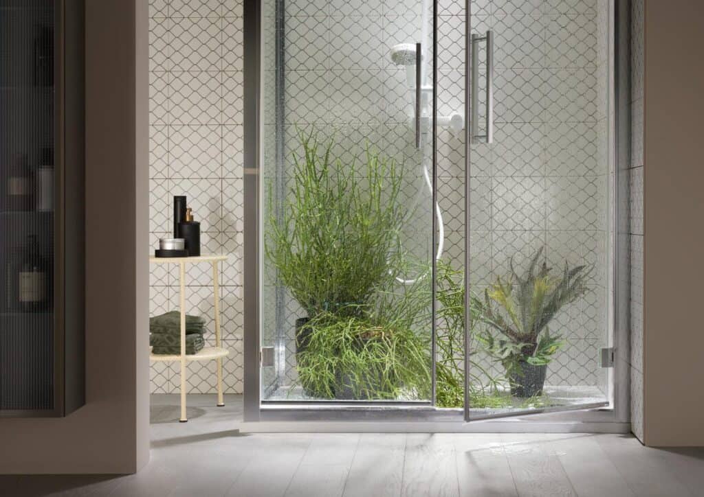 Modern bathroom with a glass shower enclosure filled with lush green plants, light wood flooring, and a small side table holding toiletries and towels.