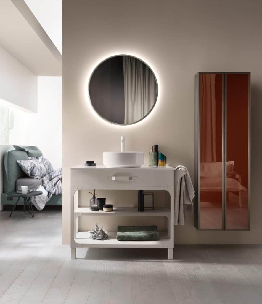 Modern bathroom with a round illuminated mirror above a white vanity, featuring a vessel sink, toiletries, and a folded towel, adjacent to a bedroom with a bed.