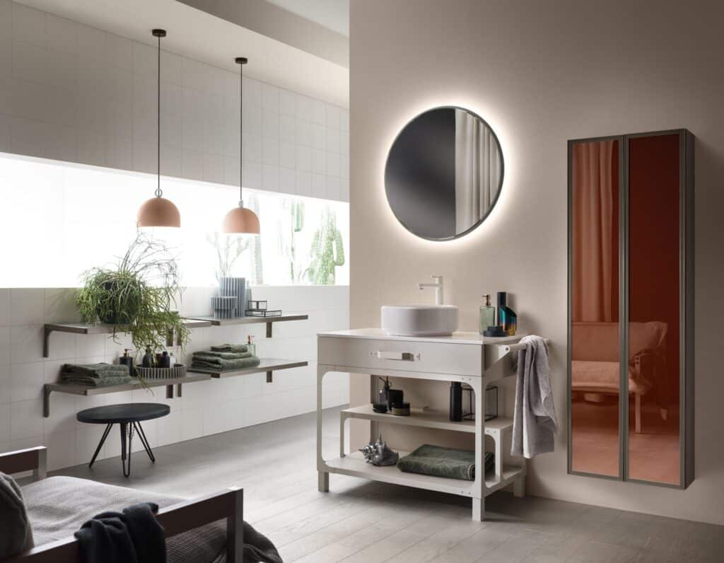 Modern bathroom interior with a round backlit mirror, minimalist sink, wall shelves with plants and towels, pendant lights, and a tall cabinet with a reflective surface.
