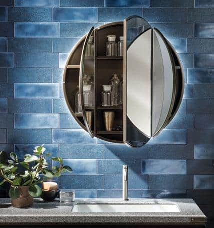 Round bathroom cabinet with mirrored doors open, revealing shelves with glass containers, set against blue tile wall above a sink and faucet.
