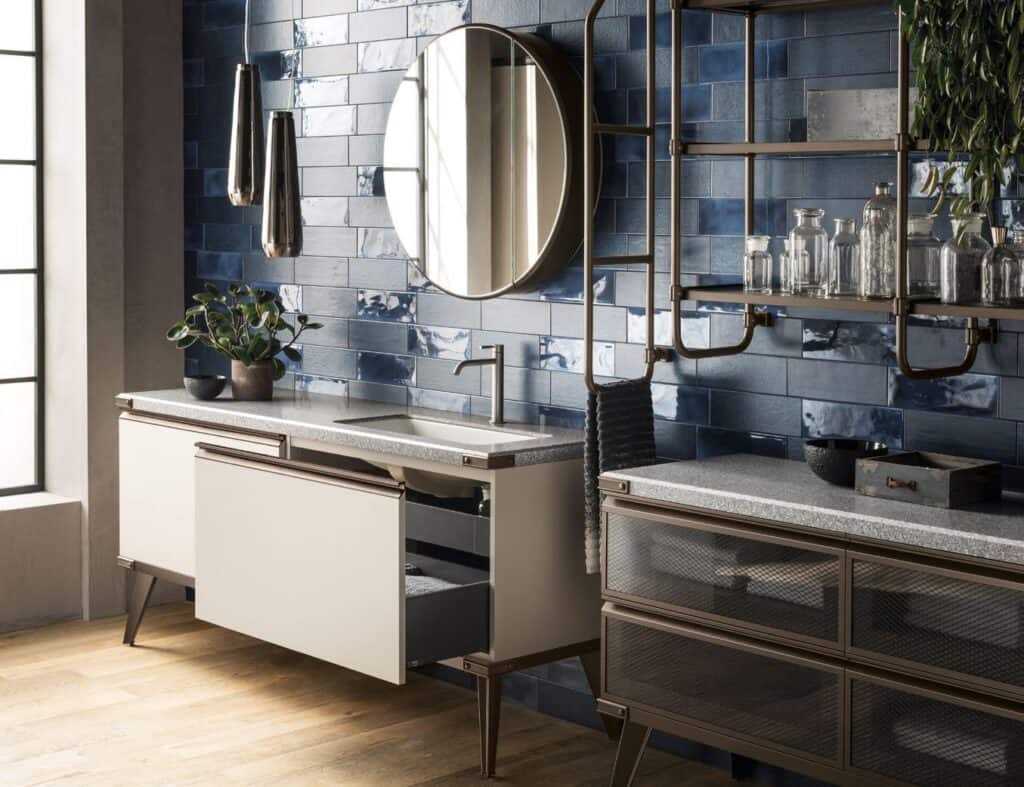 Modern bathroom with glossy blue subway tile wall, circular mirror, white vanity with open drawer, metallic pendant lights, and decorative plants and glass jars on shelving.