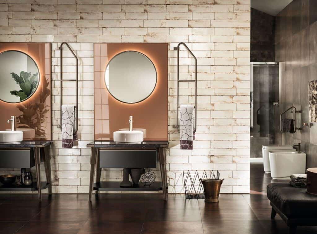 Modern bathroom with double sinks, round mirrors, industrial-style towel racks, and brick tile walls.