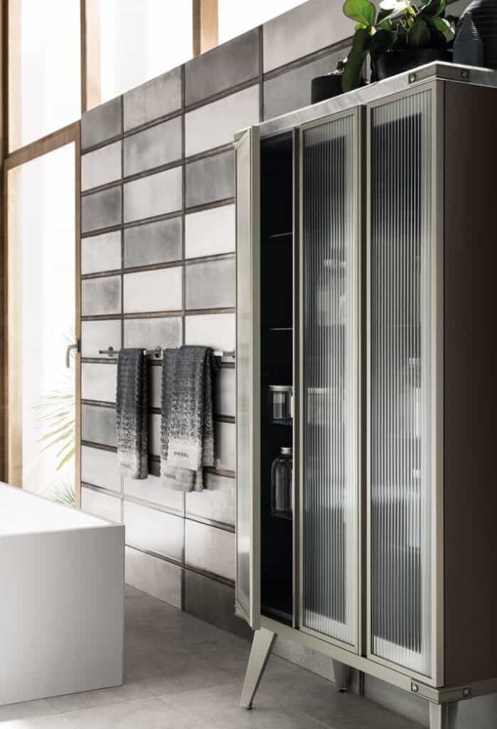 A modern bathroom with a metal cabinet featuring glass doors, a tiled wall with two hanging towels, and a white bathtub.