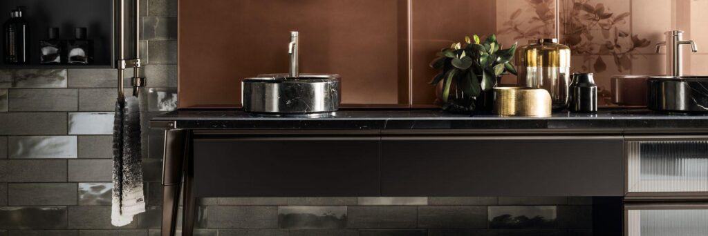 Modern bathroom with dark marble sinks, decorative jars, and leafy plant on a black countertop against a copper accent wall.