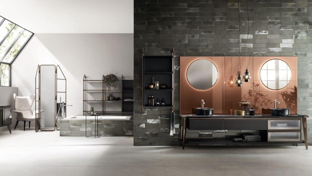 Modern bathroom interior with dark tiled walls, large vanity with dual sinks and round mirrors, a bathtub, shelving, and a chair under angled skylights.